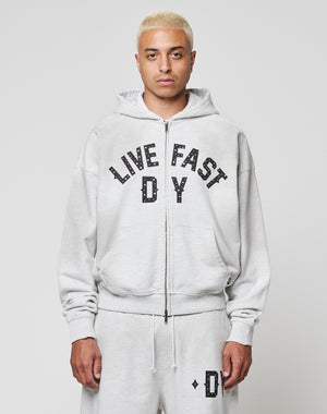 A person with short bleached blonde hair wears the LIVE FAST DIE YOUNG Live Fast Rivet Hooded Zip in light gray, styled oversized with matching sweatpants. Bold black LIVE FAST D*Y text and subtle distressing make it stand out against a white background.
