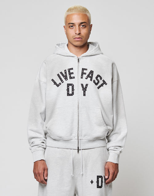 A person with short bleached blonde hair wears the LIVE FAST DIE YOUNG Live Fast Rivet Hooded Zip in light gray, styled oversized with matching sweatpants. Bold black LIVE FAST D*Y text and subtle distressing make it stand out against a white background.