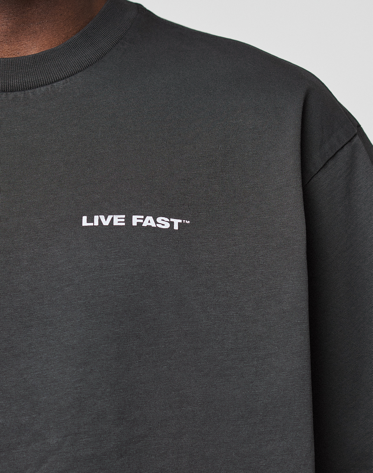 Close-up of a person in the LFDY Grand Theft Tee by LIVE FAST DIE YOUNG, featuring a dark 100% Baumwolle crew neck, LIVE FAST™ white chest print, oversize fit, and stylish front & back prints. Only part of face and shoulder shown.