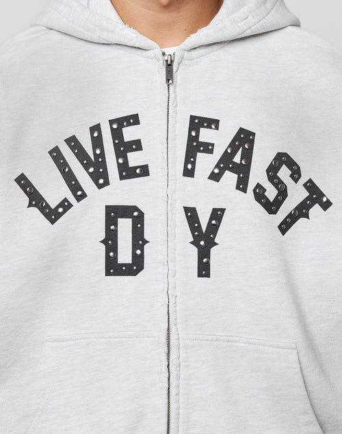 A person wears the Live Fast Rivet Hooded Zip by LIVE FAST DIE YOUNG—a light gray, oversized zip-up hoodie featuring bold black and white LIVE FAST D Y lettering across the chest. Only their lower face and upper torso are shown.