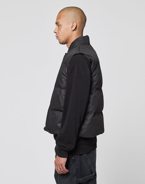 A man with a shaved head stands in profile against a plain background, wearing the LIVE FAST DIE YOUNG Basic Cropped Vest in black over a black long-sleeve shirt and dark jeans.