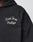 Close-up of the LFDY LÉquipe Hooded Zip by LIVE FAST DIE YOUNG—an oversized black hoodie with Live Fast Rallye embroidered in white chenille on the chest. Part of the persons neck and chin are visible; their face is largely out of frame.