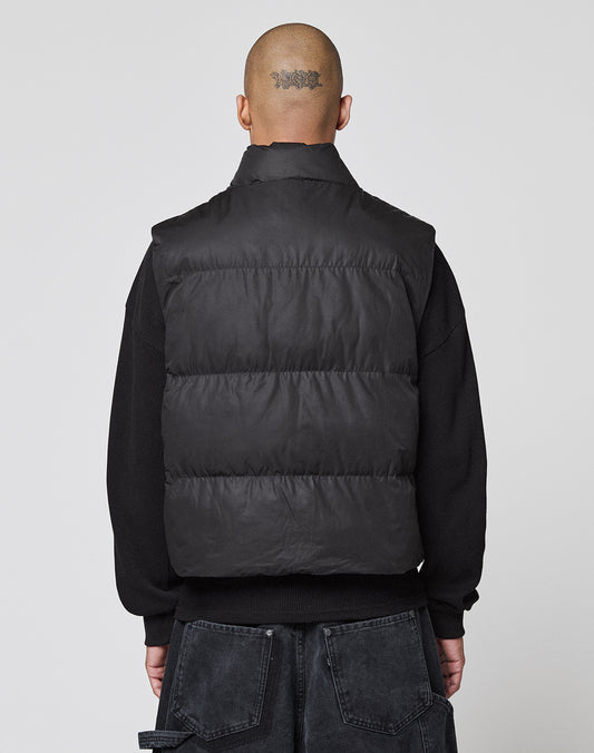 A person with a shaved head and visible tattoo faces away, wearing the LIVE FAST DIE YOUNG Basic Cropped Vest over a black long-sleeve shirt and dark cargo pants with große Taschen. The background is plain and light-colored.