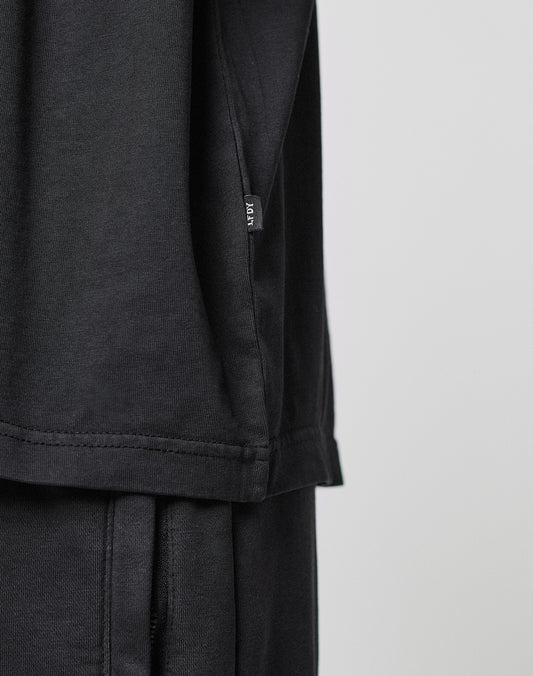 Close-up of a person wearing the LIVE FAST DIE YOUNG Basic +49 Tee, a loose-fitting black 100% Baumwolle shirt with a small black fabric tag featuring white text on the side seam. The background is plain and light-colored.