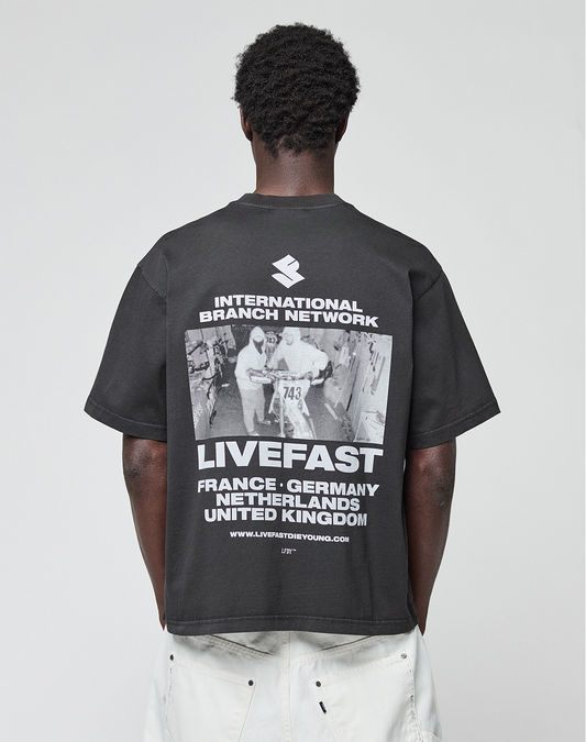 A person with short curly hair, seen from behind, wears the LIVE FAST DIE YOUNG LFDY Grand Theft Tee—an oversize black 100% cotton shirt with front and back prints: “INTERNATIONAL BRANCH NETWORK,” “LIVEFAST,” and various countries.