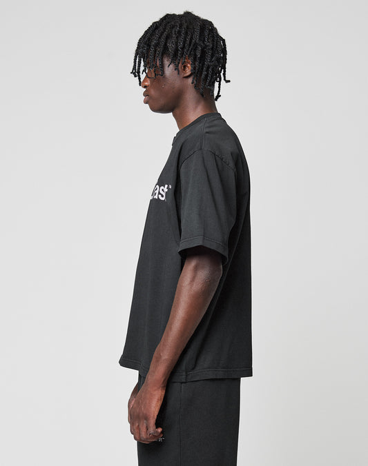 A person with short, twisted dreadlocks stands in profile against a plain light background, wearing the LIVE FAST DIE YOUNG Basic +49 Tee—an oversized black 100% Baumwolle T-shirt with a partial white frontprint text—paired with black pants.
