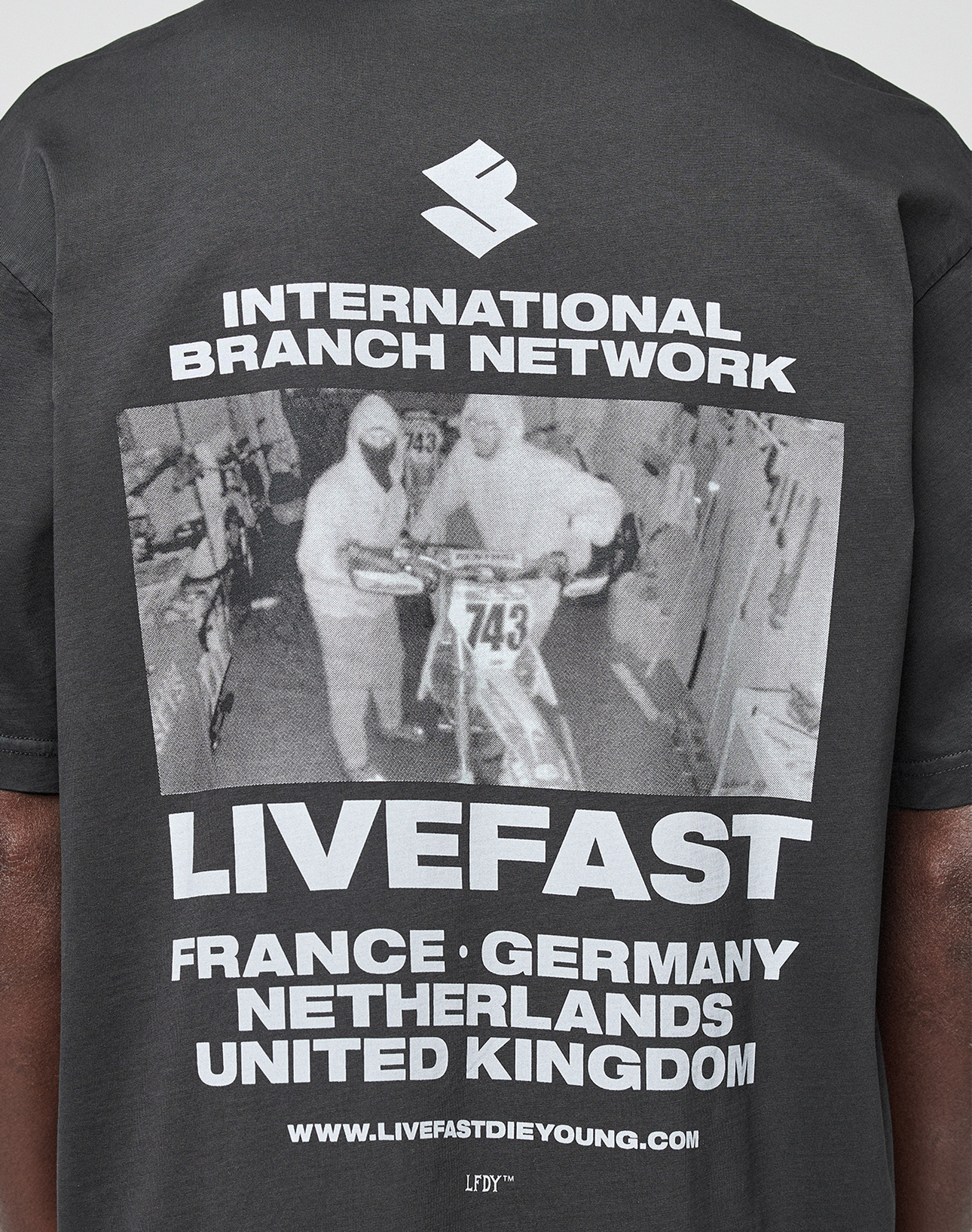 A close-up of the LFDY Grand Theft Tee by LIVE FAST DIE YOUNG shows a black-and-white image of two people with a motorcycle, “International Branch Network LIVEFAST” text, listed countries, and is made from 100% Baumwolle for everyday comfort.