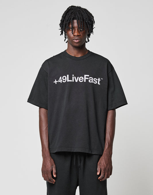 A young man with short dreadlocks wears the LIVE FAST DIE YOUNG Basic +49 Tee, an oversized black T-shirt with a bold “+49LiveFast” front print, made from 100% Baumwolle, standing against a plain light gray background.