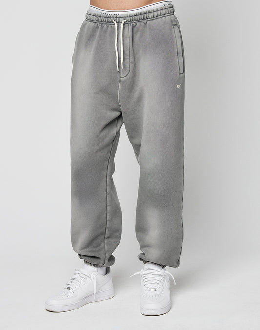 A person models the LIVE FAST DIE YOUNG Basic Loose Joggerālight gray, loose-fitting sweatpants with a logo print and white drawstringsāpaired with white sneakers against a plain light gray background.