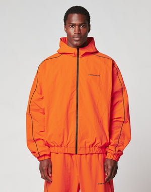 A man faces forward against a plain background, wearing the LIVE FAST DIE YOUNG Live Fast Trackjacket and matching pants in bright orange Nylon-Elastan blend, with elastic cuffs, black piping, and small text on the left chest and leg.