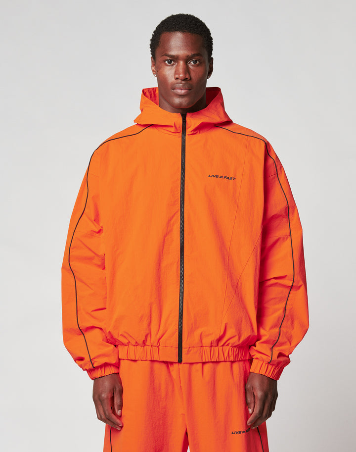 A man faces forward against a plain background, wearing the LIVE FAST DIE YOUNG Live Fast Trackjacket and matching pants in bright orange Nylon-Elastan blend, with elastic cuffs, black piping, and small text on the left chest and leg. | model