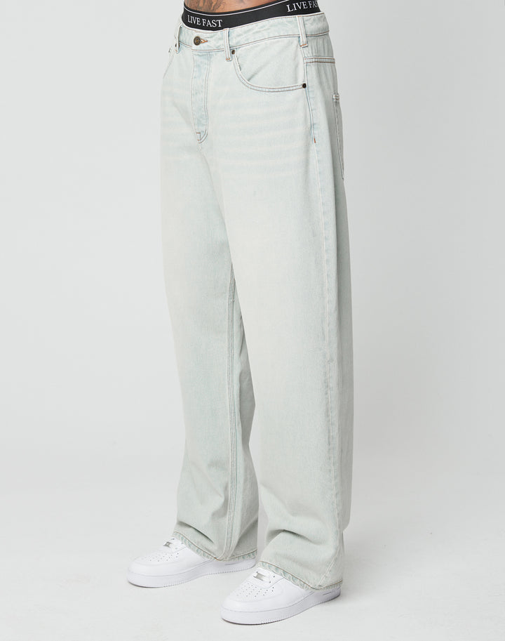 A person in LIVE FAST DIE YOUNGs LFDY Baggy Denim light-wash jeans, white sneakers, and a visible black LIVE FAST waistband stands against a plain white background.