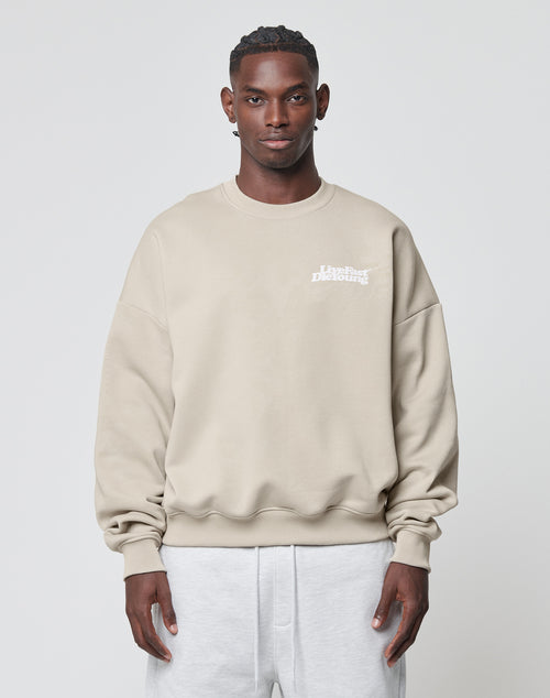 A man stands against a plain light background, wearing the LIVE FAST DIE YOUNG Basic Chest Sweater—an oversize fit beige sweatshirt with a white front print on the chest—and light gray pants, looking straight at the camera with a neutral expression.