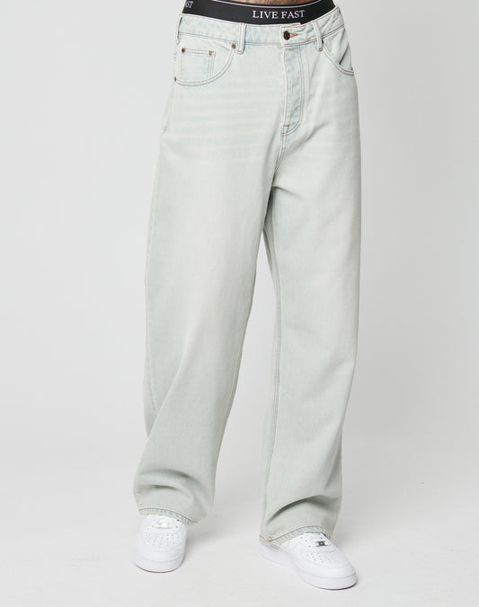 Model wears LFDY Baggy Denim by LIVE FAST DIE YOUNG—baggy fit, light-wash Baumwolle jeans with a black LIVE FAST waistband—styled simply with white sneakers against a plain white background.