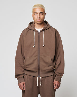 A person with short blond hair wears the LIVE FAST DIE YOUNG LFDY Lightweight Hooded Zip in brown, paired with matching pants, standing against a plain light gray background and looking directly at the camera with a neutral expression.
