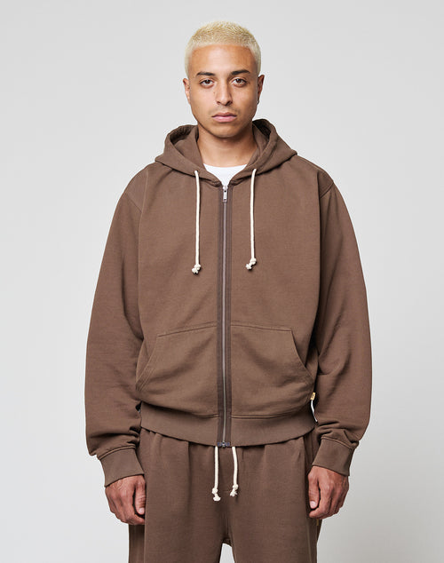A person with short blond hair wears the LIVE FAST DIE YOUNG LFDY Lightweight Hooded Zip in brown, paired with matching pants, standing against a plain light gray background and looking directly at the camera with a neutral expression.