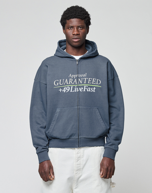 A man stands against a light gray background wearing the LFDY Guaranteed Hooded Zip by LIVE FAST DIE YOUNG, featuring an oversized dark blue fit, frontprint "Approval GUARANTEED +49 Live Fast", 100% Baumwolle. He pairs it with light-colored pants.