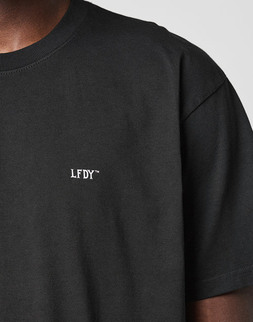 A close-up shows a person in the LIVE FAST DIE YOUNG Basic Chest Tee, a plain black regular fit Baumwolle T-shirt with a small white LFDY™ logo embroidered on the left chest. Only the upper torso and part of the neck are visible.
