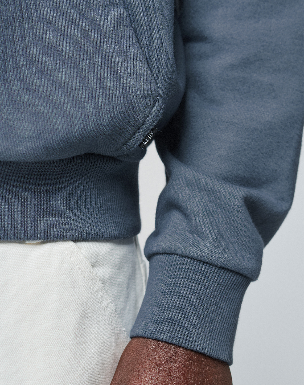 Close-up of a person wearing the blue LFDY Guaranteed Hooded Zip by LIVE FAST DIE YOUNG, paired with off-white pants. Part of their hand and arm is visible against a neutral background.