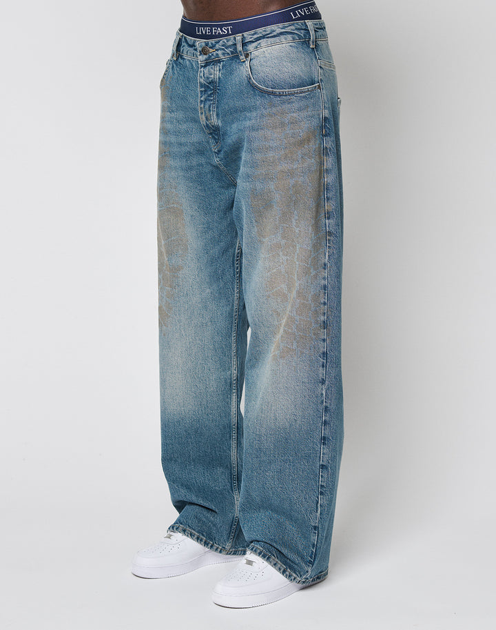Someone wearing LIVE FAST DIE YOUNG LFDY Tire Tracks Denim, baggy fit, light blue, slightly faded 100% Baumwolle jeans with a high waist, white sneakers, and black LIVE FAST waistband underwear against a plain white background.