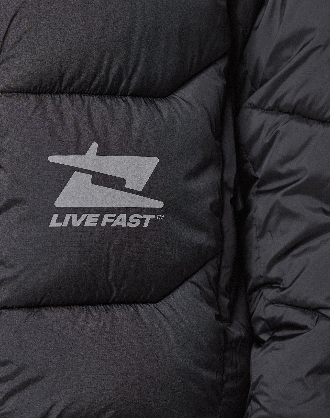Close-up of the Live Fast Puffer Jacket by LIVE FAST DIE YOUNG, a black quilted jacket with a stylized jet logo and LIVE FAST™ in white reflektierender Print for enhanced visibility.