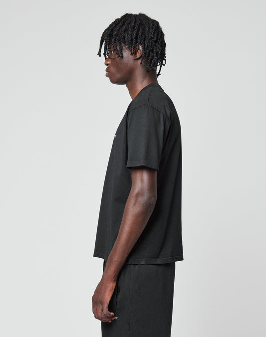 A person with short, twisted hair stands in profile against a plain background, wearing the LIVE FAST DIE YOUNG Basic Chest Tee—a regular-fit black T-shirt with a subtle logo print—paired with matching black Baumwolle pants.