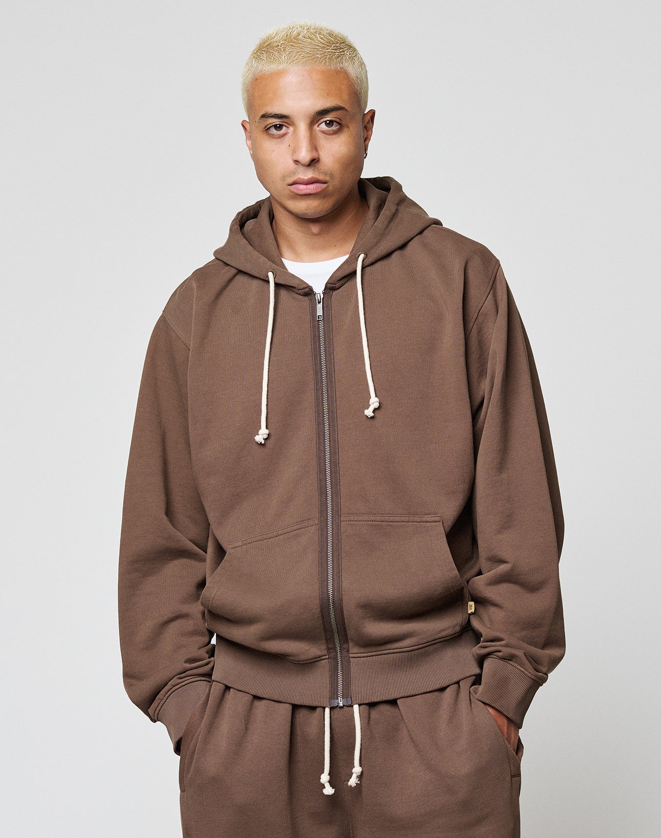 A person with short, bleached blond hair wears the LIVE FAST DIE YOUNG LFDY Lightweight Hooded Zip in brown with cream drawstrings and matching pants, standing against a plain light gray background.