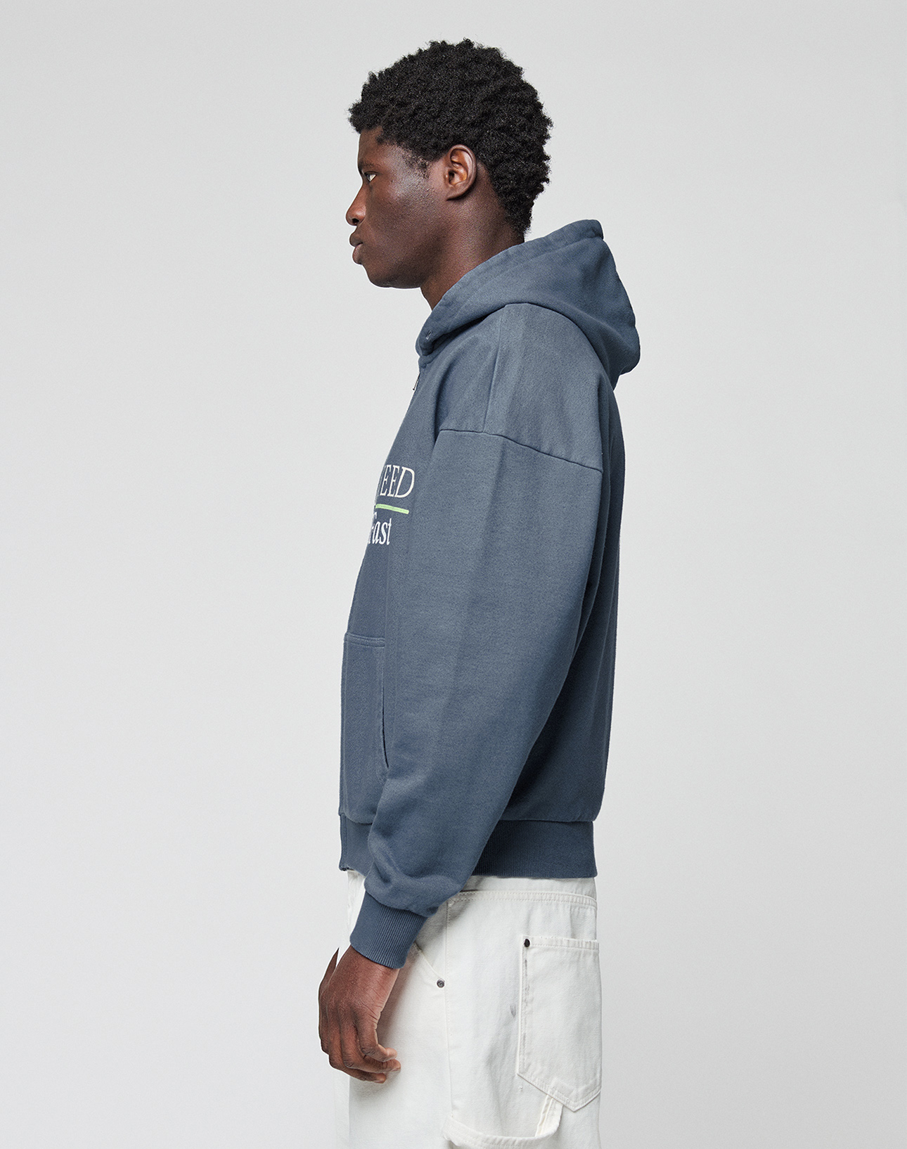 A person with dark curly hair stands in profile, wearing the LIVE FAST DIE YOUNG LFDY Guaranteed Hooded Zip—an oversized blue hoodie with white front print—and loose white pants, posed against a plain light background.