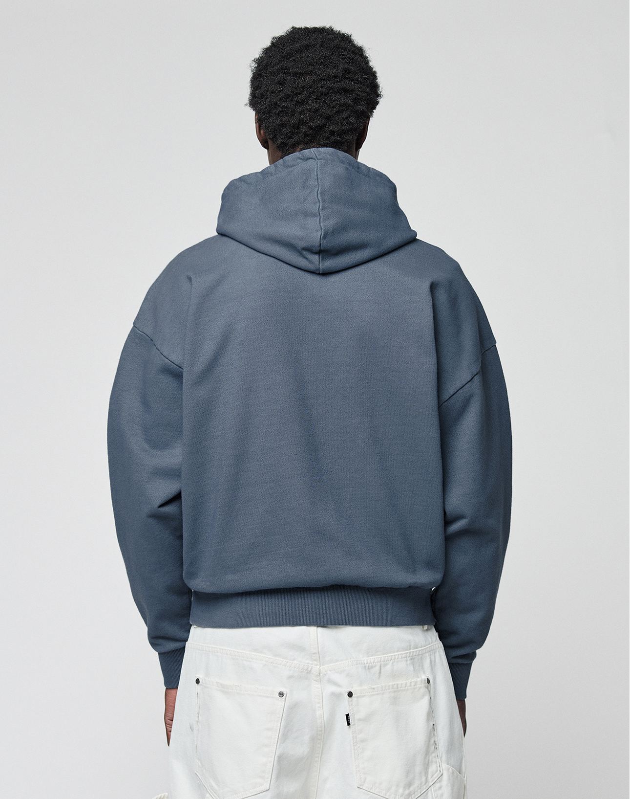 A person with short curly hair stands with their back to the camera in the LIVE FAST DIE YOUNG LFDY Guaranteed Hooded Zip, an oversize-fit blue hoodie made from 100% Baumwolle, styled with white pants featuring visible back pockets against a plain light background.