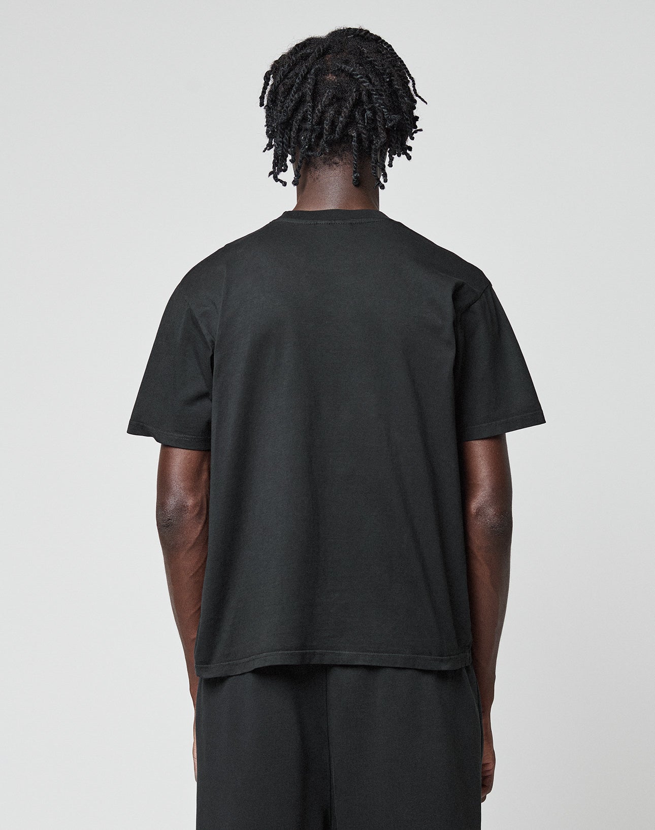 A person with short, twisted hair stands facing away from the camera, wearing the LIVE FAST DIE YOUNG Basic Chest Tee in black and matching pants. The regular fit tee is crafted from hochwertige Baumwolle for comfort and style against a light gray background.