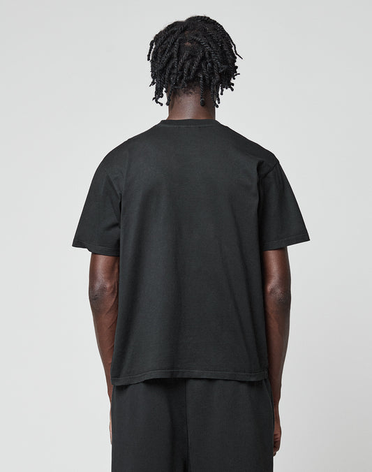 A person with short, twisted hair stands facing away from the camera, wearing the LIVE FAST DIE YOUNG Basic Chest Tee in black and matching pants. The regular fit tee is crafted from hochwertige Baumwolle for comfort and style against a light gray background.