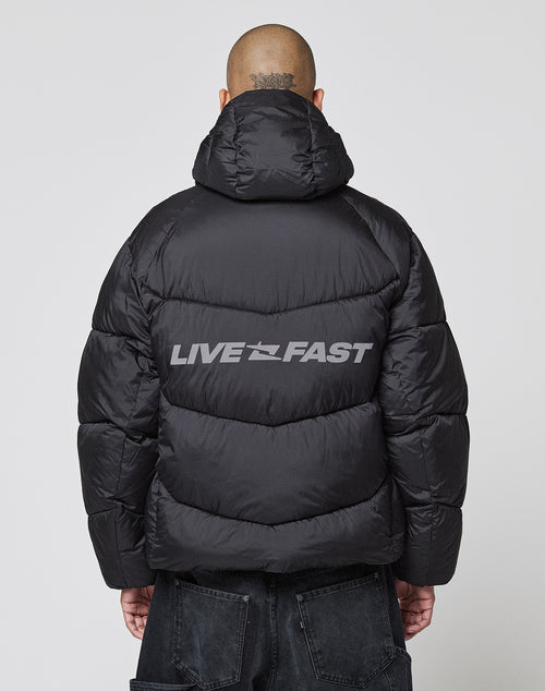A person with a shaved head and neck tattoo is seen from behind wearing the LIVE FAST DIE YOUNG Live Fast Puffer Jacket, an oversized black puffer featuring “LIVE FAST” in bold light gray letters and a reflective print on the back.