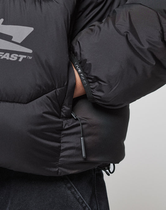 Close-up of a person in the LIVE FAST DIE YOUNG Live Fast Puffer Jacket, black with an oversize fit, visible zipper pocket, and reflective FAST logo print. The hand is tucked into a side pocket, adding a modern touch.