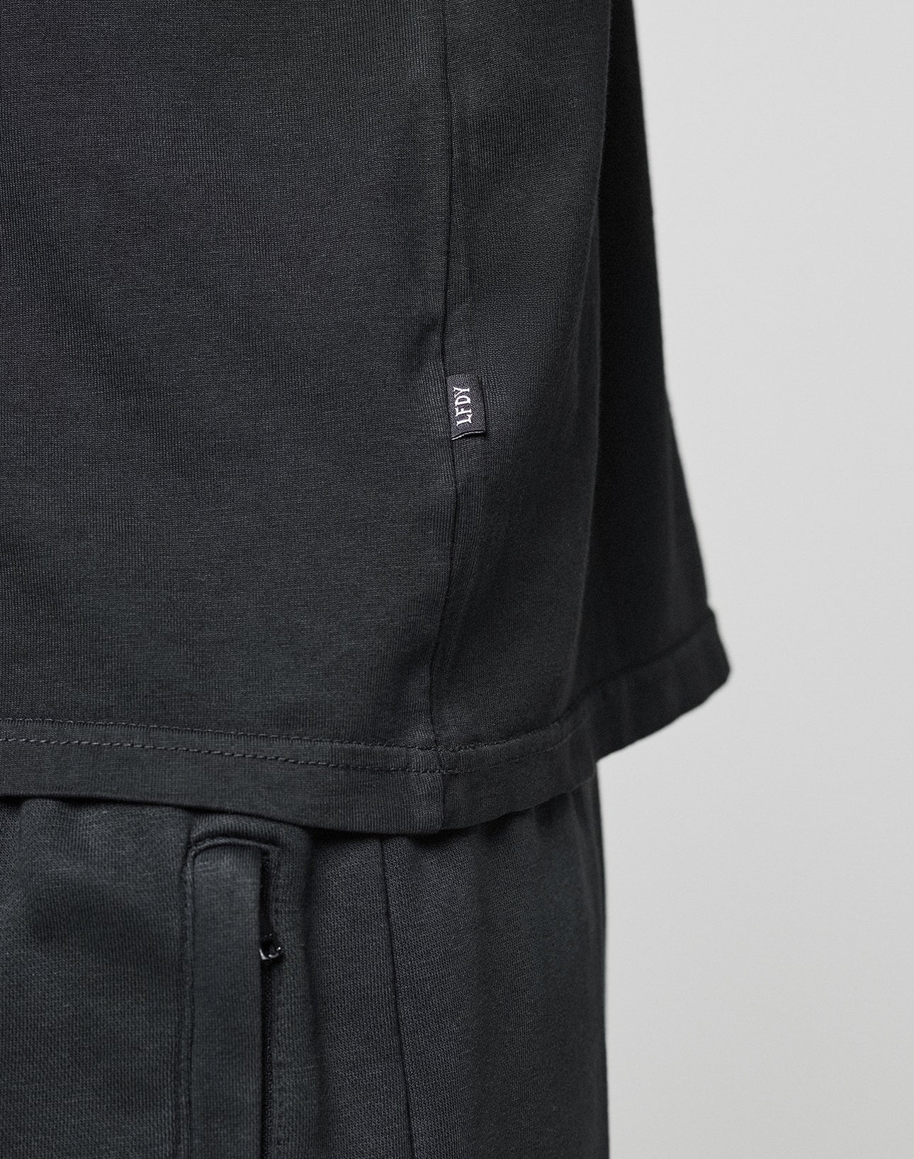 Close-up of a person wearing the LIVE FAST DIE YOUNG Basic Chest Tee in black and matching pants. A small fabric tag with white text is sewn into the side seam. The background is plain and light-colored.