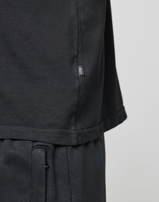 Close-up of a person wearing the LIVE FAST DIE YOUNG Basic Chest Tee in black and matching pants. A small fabric tag with white text is sewn into the side seam. The background is plain and light-colored.