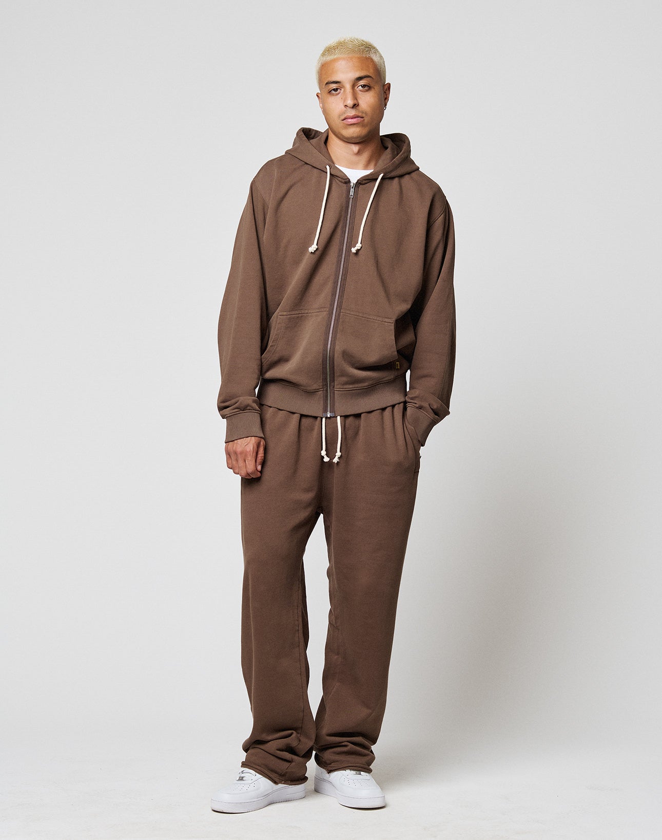 A person with short blond hair wears the LIVE FAST DIE YOUNG LFDY Lightweight Hooded Zip and matching sweatpants made from 100% cotton, paired with white sneakers, standing against a plain light background.
