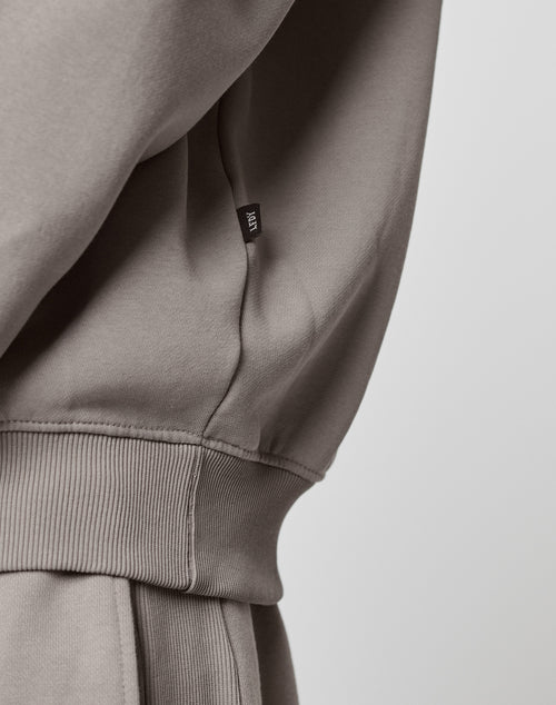 Close-up of the LIVE FAST DIE YOUNG Basic Blank Sweater in taupe Oversize Fit, featuring a ribbed waistband and a small black side tag, set against a plain light background.