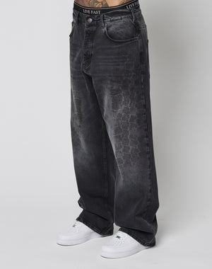Person wearing LIVE FAST DIE YOUNG’s LFDY Tire Tracks Denim—baggy, loose-fitting, faded black jeans with a subtle tire track texture and black branded waistband—paired with white sneakers against a plain white background.