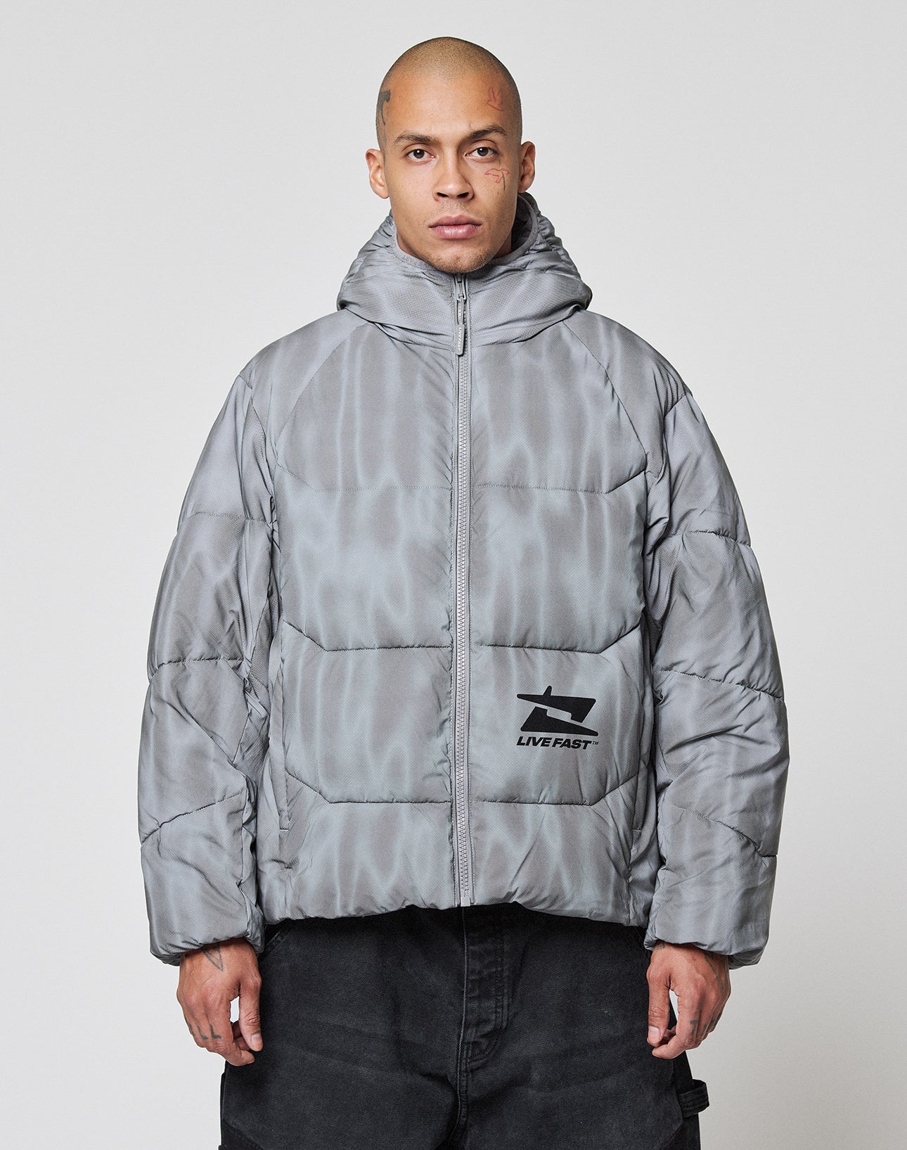 A person with a shaved head wears the LIVE FAST DIE YOUNG Live Fast Puffer Jacket, an oversized, shiny gray hooded jacket with a reflective finish and graphic logo on the lower left. The background is plain and light-colored.