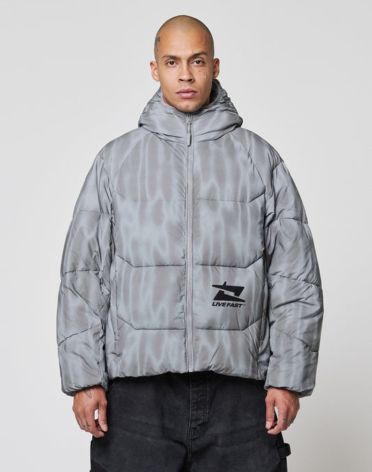 A person with a shaved head wears the LIVE FAST DIE YOUNG Live Fast Puffer Jacket, an oversized, shiny gray hooded jacket with a reflective finish and graphic logo on the lower left. The background is plain and light-colored.
