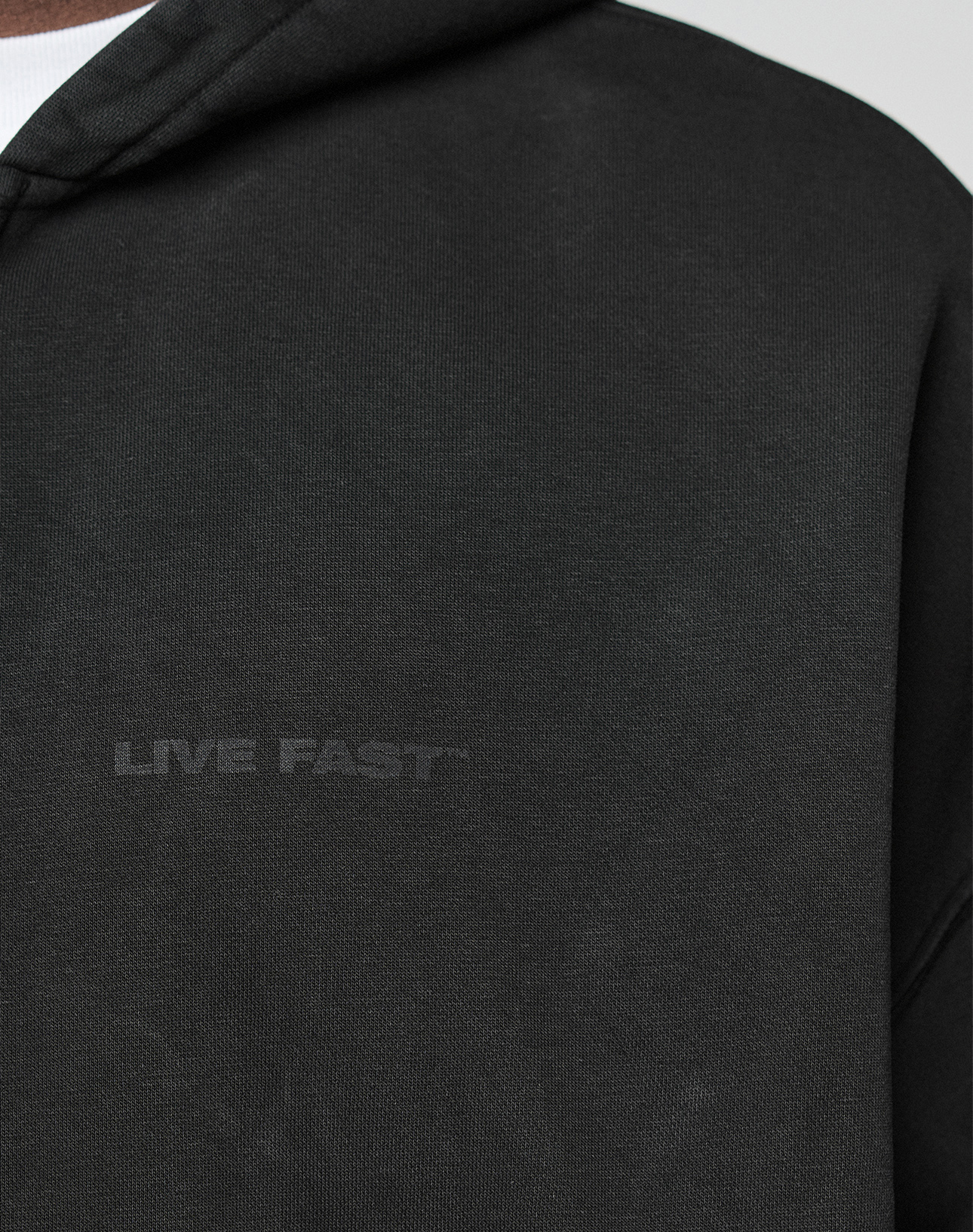 Close-up of the black LFDY Grand Theft Hooded Zip by LIVE FAST DIE YOUNG in Oversize Fit, featuring subtle dark LIVE FAST lettering on the chest. The hood and part of a white shirt underneath are visible.
