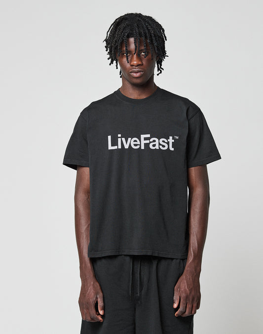 A person with short, twisted hair models the Basic LiveFast Tee by LIVE FAST DIE YOUNG—a regular fit black t-shirt with a bold white LiveFast™ front print, showcased against a plain light gray background.