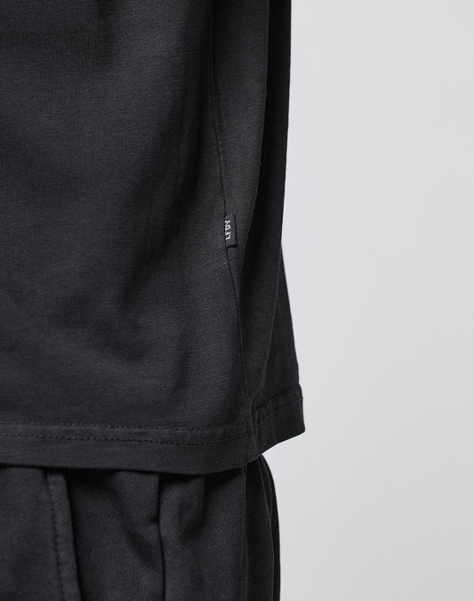 Close-up of a person wearing the LIVE FAST DIE YOUNG Basic LiveFast Tee in black and black pants, featuring a small black tag near the shirt’s side seam against a plain, light background.