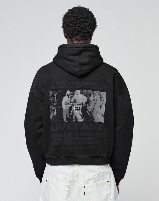 A person wears the LFDY Grand Theft Hooded Zip by LIVE FAST DIE YOUNG, an oversize fit black hoodie with Kapuze, grayscale runner photo, “LIVE FAST” text, and country names. Made from soft Baumwolle for all-day comfort.