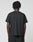 A person with short, twisted hair stands with their back to the camera, wearing a plain black Basic LiveFast Tee by LIVE FAST DIE YOUNG and black pants against a light gray background.
