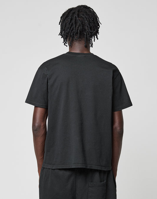 A person with short, twisted hair stands with their back to the camera, wearing a plain black Basic LiveFast Tee by LIVE FAST DIE YOUNG and black pants against a light gray background.