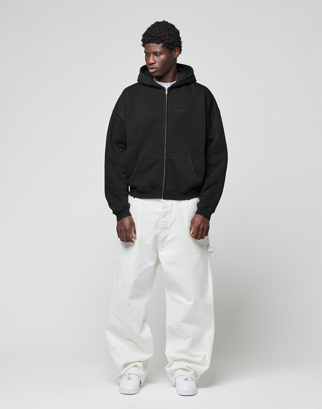 A person stands against a plain background wearing the LIVE FAST DIE YOUNG LFDY Grand Theft Hooded Zip in black, loose oversized white pants, and white sneakers, looking slightly to the side with hands near their pockets.