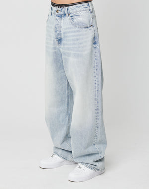 Person trägt die LFDY Baggy Denim von LIVE FAST DIE YOUNG aus 100% Baumwolle in Light-Wash-Optik mit Faded-Effekt und weitem, geradem Bein zu weißen Sneakern vor weißem Hintergrund. High-Waist-Schnitt, lockere Passform. Oberkörper nicht sichtbar.