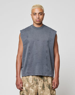 A person with short bleached blonde hair wears the LIVE FAST DIE YOUNG Heavy Muscle Tank, a sleeveless faded gray top with a bold front print, paired with brown camo cargo shorts, standing against a plain white background.