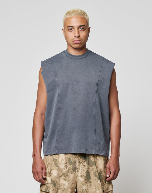 A person with short bleached blonde hair wears the LIVE FAST DIE YOUNG Heavy Muscle Tank, a sleeveless faded gray top with a bold front print, paired with brown camo cargo shorts, standing against a plain white background.